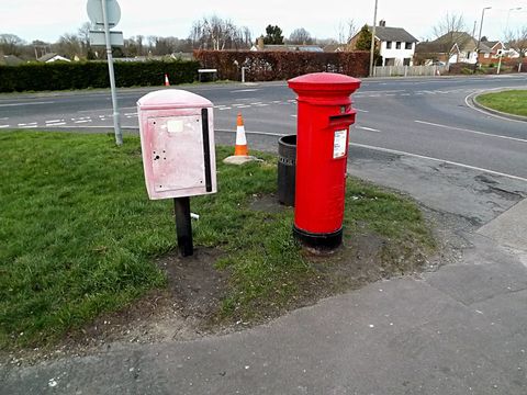 Red Royal Mail pillar box beside a faded grey storage box at a road junction