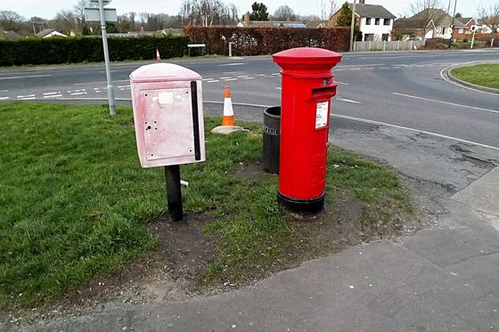 Red Royal Mail pillar box beside a faded grey storage box at a road junction
