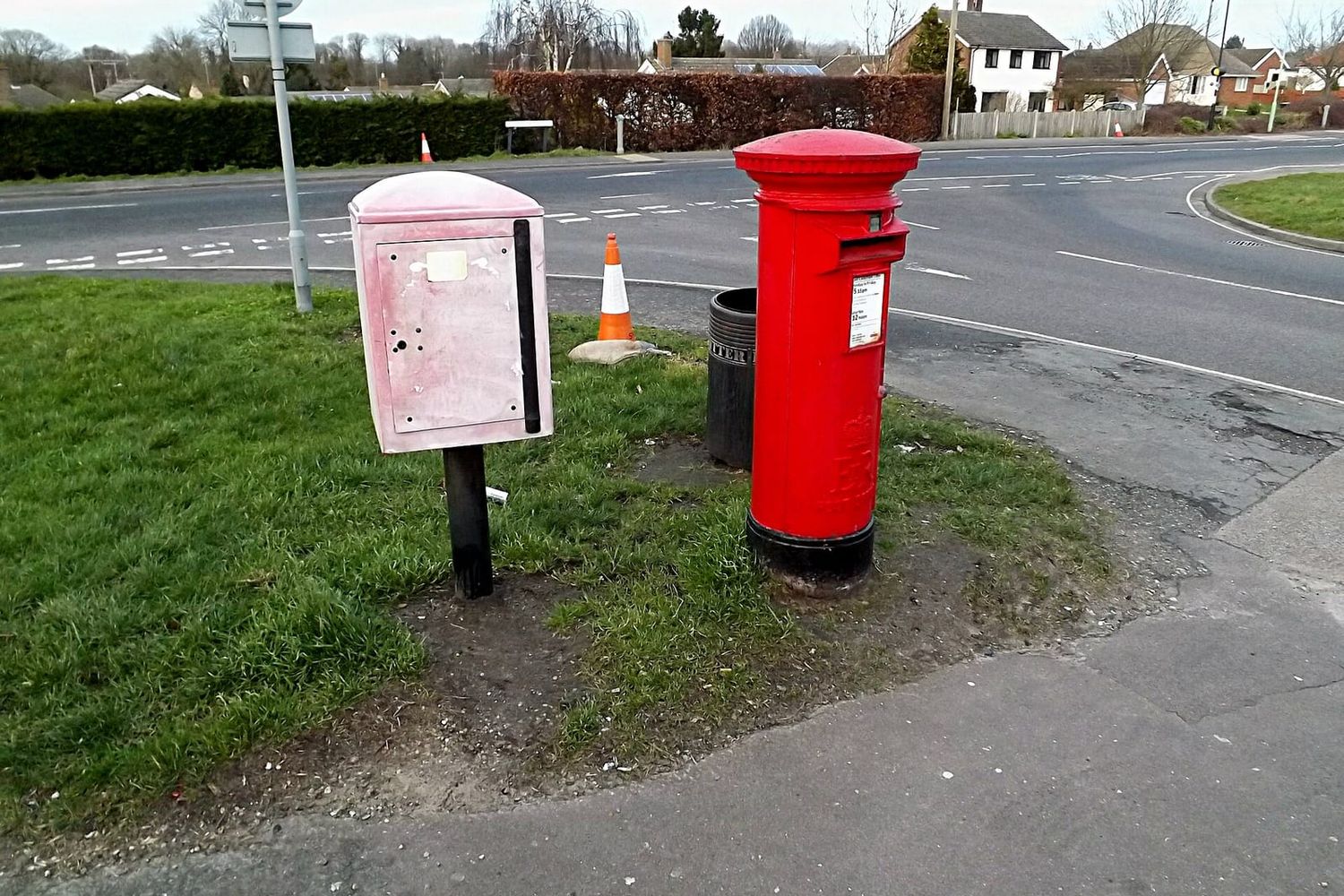 Red Royal Mail pillar box beside a faded grey storage box at a road junction